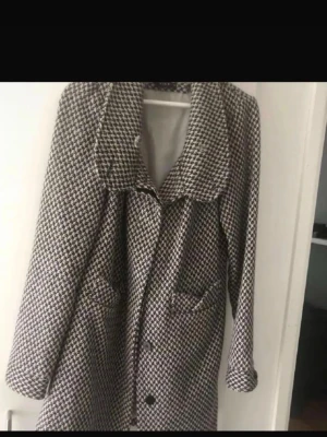 Rutig kappa med stora slag - Snygg rutig kappa i svart och vitt med stora slag och knappar framtill. Jackan har två fickor och är lång i modellen, perfekt för dig som gillar klassisk stil med en twist. Materialet känns mjukt och passar bra till kyligare dagar.