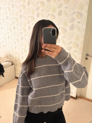 Grå randig stickad tröja - Säljer en mysig grå stickad tröja med vita ränder. Tröjan har en croppad passform, ribbade muddar och lång ärm. I storlek 164