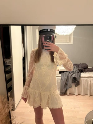Vit spetsklänning med volang - Superfin kort vit spetsklänning med långärmade transparenta ärmar och blommigt mönster. Klänningen har volang nertill och är figurnära i modellen. Perfekt till student eller sommarens alla tillfällen! Endast använd en gång