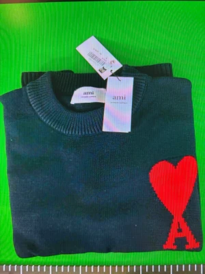 Svart stickad tröja från Ami - Snygg svart stickad tröja från Ami med rund halsringning och ribbade muddar. Tröjan har en stor röd hjärtapplikation med bokstaven A på bröstet, vilket ger en unik och trendig look. Perfekt för dig som vill ha något stilrent men ändå sticker ut.