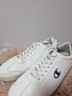 Vita sneakers från Champion strl 44 - Säljer ett par klassiska vita sneakers från Champion i storlek 44. Skorna har en låg profil, rund tå och snörning framtill. Tillverkade i syntetmaterial med en stilren design och mörkblå Champion-logga på sidan. Perfekta för en clean och sportig look.