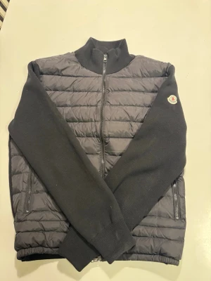 Moncler cardigan svart - Säljer en svart cardigan från Moncler med quiltad front och stickade ärmar. Jackan har hög krage, två dragkedjeförsedda fickor och hel dragkedja framtill. Klassisk Moncler-logga på ena ärmen. Perfekt för dig som vill ha en stilren och bekväm jacka. Jackan passar mig som är runt 180cm och väger cirka 75kg. Skriv till mig för mer info. Jackan är väldigt lätt använd, köpte den begagnad för ett par månader sedan men har knappast använt den på grund av kylan. Den har inga fläckar eller hål.