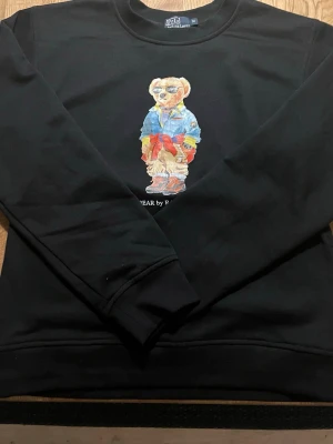 Svart sweatshirt Polo Bear Ralph Lauren - Svart sweatshirt från Polo Ralph Lauren med ikoniskt Polo Bear-tryck framtill. Tröjan är i 100% bomull och har rund hals samt långa ärmar. Perfekt statement-plagg för dig som gillar streetstyle och klassiska märken. Skriv fråga vid minsta fundering 🤔. Pris kan alltid diskuteras vid snabb affär🚚♻️