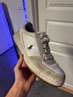 Vita sneakers från Polo Ralph Lauren - Snygga vita sneakers från Polo Ralph Lauren med klassisk logga på sidan. Skorna har grå mockadetaljer, snörning och en brun sula med tydligt Polo-märke undertill. Perfekta till jeans eller shorts för en clean och sportig look.