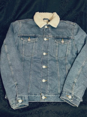 Jeansjacka med teddykrage, stl S - Säljer en klassisk blå jeansjacka från Weekday med vit teddykrage. Jackan har raka sömmar, två bröstfickor med knappar och stängs med silverfärgade knappar framtill. Perfekt för dig som vill ha en chill och trendig look.