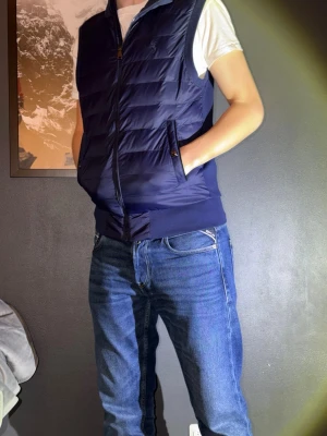 Mörkblå dunväst från Polo Ralph Lauren - Säljer en mörkblå dunväst från Polo Ralph Lauren med klassisk broderad logga på bröstet. Västen har quiltad design, dragkedja framtill och två fickor med knappstängning. Perfekt att slänga över en hoodie eller t-shirt för en clean och sportig look. Helt oanväd. Ny pris runt 2400. Han på bilden är 177 och storlek på västen är XS