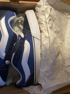 Blå och vita Vans Old Skool sneakers - Säljer ett par klassiska Vans Old Skool sneakers i marinblått och vitt. Skorna har den ikoniska vita sidan, vit sula och svart detalj runt sulan. Ovandelen är i mocka och canvas med vita sömmar och snörning. Perfekta för dig som gillar streetwear och skate-stil.