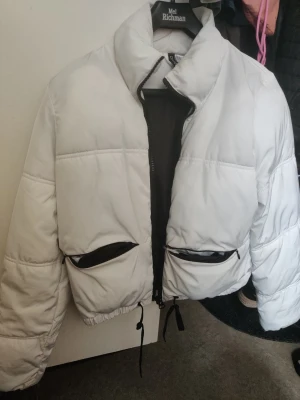 Vit pufferjacka från H&M Divided XS - Säljer en vit pufferjacka från H&M Divided i storlek XS. Jackan har svarta dragkedjor, två stora fickor framtill och snörning i nederkant. Perfekt för kyliga dagar och ger en cool, oversized vibe.