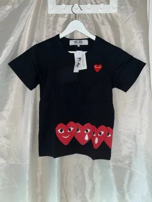 CDG t-shirt - Helt ny t-shirt från Comme des Garçons Play med korta ärmar och rund hals. Framtill finns ett rött mindre hjärta och flera tryckta hjärtan med olika ansiktsuttryck längs nederkanten som fortsätter baktill. Axelbredd 39cm, längd 61cm. 1,22