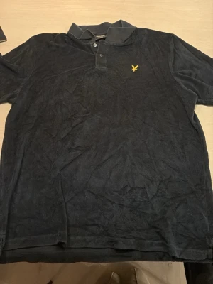 Mörkblå piké i frotté material från Lyle & Scott - Säljer en mörkblå henley från Lyle & Scott i frotté . Skjortan har klassisk krage, två knappar framtill och den ikoniska gula loggan broderad på bröstet. Relaxed fit för en chill vibe.