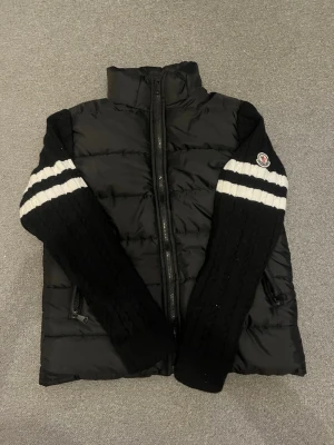 Svart Moncler pufferjacka med stickade ärmar - Säljer en svart pufferjacka från Moncler med stickade, kabelmönstrade ärmar och vita ränder. Jackan har dragkedja framtill, två sidofickor med dragkedja och Moncler-logga på ena ärmen. Perfekt för dig som vill ha en unik och sportig look.