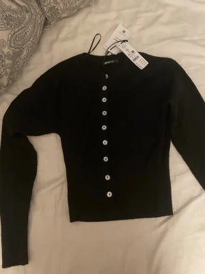 Svart kofta från Gina Tricot - Säljer en svart kofta från Gina Tricot med långa ärmar och vita knappar hela vägen framtill. Koftan är figurnära. Helt ny och aldrig använd 