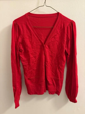 Röd kofta med puffärm och knappar - Röd kofta med v-ringning, puffärm och knäppning framtill. Tröjan har två små fickor framtill och är ribbad vid ärmslut och nederkant. Tillverkad i mjuk bomull, perfekt för lager på lager under höst och vinter.