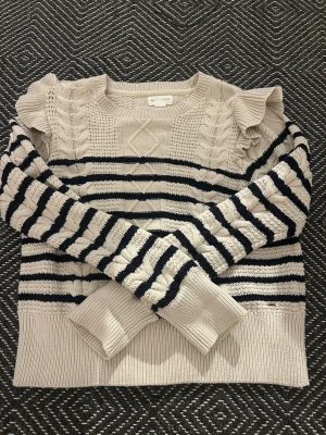 Randig stickad tröja med volang - Supersöt beige och svart stickad tröja från Holly and white med breda ränder, flätstickade detaljer och volang vid axlarna. Tröjan har lång ärm och ribbade muddar. Perfekt för dig som vill ha en mysig men ändå stylish look.