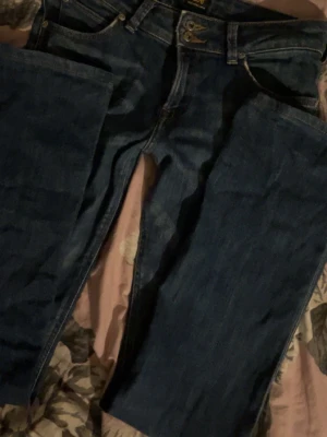 Mörkblå Lee jeans bootcut - Säljer ett par mörkblå jeans från Lee med klassisk femficksdesign och bootcut-ben. Jeansen har snygga kontrastsömmar och ett brunt Lee-läderpatch bak i midjan. Tillverkade i slitstarkt denimtyg som ger en tidlös look. Storlek W27 L31. 