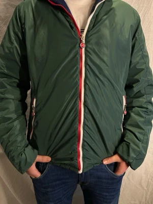Grön vindjacka från Colmar - Snygg grön vindjacka från Colmar med huva och dragkedja i vitt och rött. Jackan har två fickor med dragkedja och en broderad Colmar-logga på ärmen. Perfekt för dig som vill ha en stilren och sportig look. Jackan är vindtålig samtidigt som den är lite varmare 
