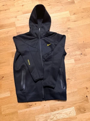 Nike NOCTA svarta mjukisbyxor - Svarta mjukisbyxor från Nike NOCTA med gul logga och detaljer. Byxorna har dragsko i midjan, raka ben och två fickor med dragkedja. Tillverkade i mjukt material som känns skönt mot huden. Snygga broderade NOCTA- och Nike-detaljer i gult på benet.