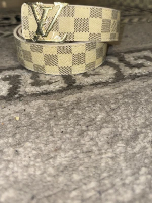 Rutig beige bälte - Säljer ett snyggt bälte i beige och vitt med klassiskt rutigt mönster och stor -logga i guld på spännet. Perfekt accessoar för att lyfta vilken outfit som helst.