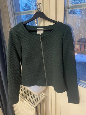 Mörkgrön cardigan jacka från Vila - Snygg mörkgrön cardigan jacka från Vila i storlek L. Jackan har ett diskret mönster, rund halsringning och dragkedja framtill. Materialet är mjukt och känns lite stickat, perfekt för dig som gillar stilrena och bekväma plagg. Det står storlek L men den passar M-S