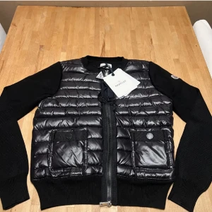 Svart quiltad pufferjacka från Moncler - Säljer en svart pufferjacka från Moncler med quiltad framsida och två stora fickor med knapp. Jackan har stickade ärmar och rygg, rund halsringning och dragkedja framtill. Perfekt för dig som vill ha en stilren och exklusiv look.