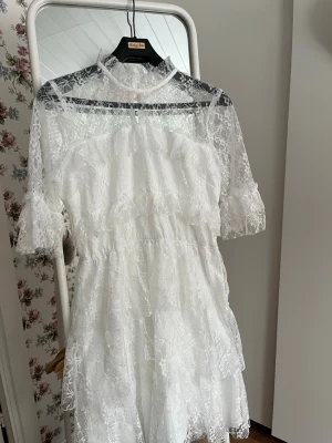Vit spetsklänning med volanger - Supersöt vit kortklänning i spets med volangdetaljer och hög hals. Klänningen har genombrutna spetsärmar och flera lager. Perfekt för sommarens alla tillfällen och studentfirande. Klänningen har lite smink vid kragen, men går bort vid tvätt.