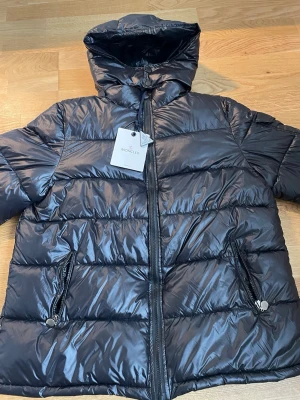 Svart Moncler pufferjacka med huva - Säljer en svart pufferjacka från Moncler med glansig finish och huva. Jackan har dragkedja framtill, två snedställda fickor med dragkedja och Moncler-logga på ärmen. Perfekt för kalla dagar och riktigt snygg till streetwear-stilen.