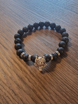 Armband med vargmotiv - Armband med lavastenpärlor och en detalj i form av ett vargmotiv. 19 cm omkrets. Pärlorna 0.8 cm. Perfekt för att komplettera din stil.