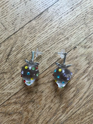 Glittriga jordgubbsörhängen med stenar - Säljer ett par unika örhängen formade som jordgubbar i transparent plast med färgglada glittriga stenar i rött, blått, gult, grönt och lila. Örhängena är ca 3,5 cm långa och har silvriga detaljer upptill. Perfekta för dig som vill sticka ut!