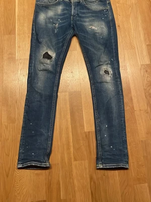 Blå slitna skinny jeans med hål - Säljer ett par mörkblåa dondups med slitningar på knäna och coola färgstänk. Jeansen har en tight passform och klassisk femficksdesign. Perfekta för dig som gillar en edgy och avslappnad stil. Nyköpta i bytik 4,5k