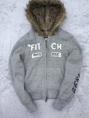 Abercrombie & Fitch – Hoodie från 1892dragkedja och pälsfodrad blus medpälsmönster - En varm och snygg Abercrombie & Fitch-tröja i en ljusgrå nyans 🤍. Med en huva med mjukt pälsfoder och fuskpäls kant ger den exceptionell komfort på kallare dagar ❄️. Broderade framtill är orden “FIT CH” och “PHYS EDUC” och en dekorativ “1892”- applikation på ärmen, som refererar till märkets grundår. Insidan är fodrad med tjockt, mysigt “fårskinn”, perfekt för vintermånaderna 🐑.  📏 Mått ✓ Total längd: 53 cm ✓ Ärmahålsbredd: 44 cm ✓ Etikettstorlek: M passar damstorlek S  📌 Skick Tröjan är i mycket