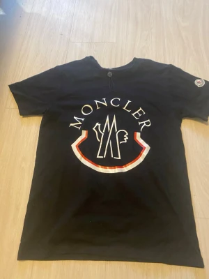 Svart Moncler t-shirt med stor logga - Svart t-shirt från Moncler med stor vit och röd logga framtill och klassisk patch på ärmen. T-shirten har rund hals, korta ärmar och är gjord i mjuk bomull. Perfekt för dig som vill ha en clean men ändå statement look.