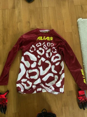 Röd och vit tröja med leopardmönster - Säljer en långärmad tröja från Alias i rött och vitt med leopardmönster och gul logga på bröstet och ärmen. Perfekt för dig som gillar sportig och unik stil. Materialet känns lätt och andas bra.