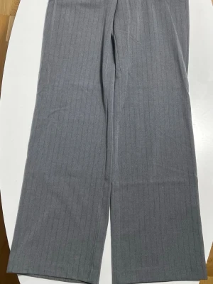 Grå randiga kostymbyxor Stradivarius - Säljer ett par grå kostymbyxor från Stradivarius i storlek 36. Byxorna har diskreta vertikala ränder, hög midja och vida ben. Klassisk look med hällor för bälte och stängs med knapp och dragkedja. Perfekta för en clean och stilren outfit.