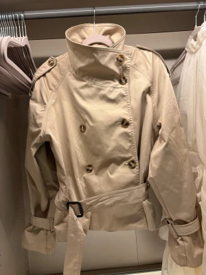 Kort beige trenchcoat från H&M - Säljer en kort trenchcoat från H&M i storlek S. Jackan är varsamt använd och i mycket fint skick, med minimala spår av användning.            Modellen är mycket eftertraktad och har varit slutsåld länge, vilket också återspeglas i priset.  Självklart skickas jackan nytvättad och fräsch.