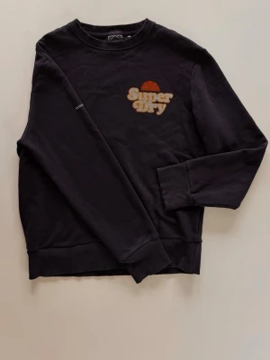 Mörkblå sweatshirt från Superdry - Mörkblå sweatshirt från Superdry med retroinspirerad logga i orange och gul på bröstet. Tröjan har rund hals, långa ärmar och ribbade muddar vid ärmslut och nederkant. Tillverkad i mjuk bomull för skön vardagsstil.                            Använd någon enstaka gång
