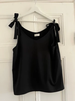 Svart linnetopp med knyt från Norr - Säljer denna silkesaktig topp från NORR med breda axelband som knyts i rosetter på axlarna. Toppen har en avslappnad passform och är tillverkad i ett mjukt, slätt material. Tveka inte på att höra av dig om du har frågor❤️