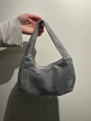 Glittrig silver axelväska - Säljer en glittrig axelväska i silver med ett tätt nät av små paljetter som ger ett riktigt statement-utseende. Väskan har en mjuk, rektangulär form och stängs med dragkedja upptill. Perfekt för dig som vill sticka ut och addera lite extra bling till din outfit.