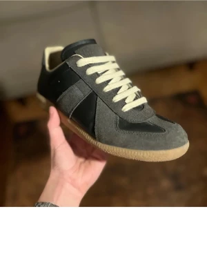 Maison margiela  - Säljer ett par stilrena sneakers i grå mocka och svart skinn med vita snören och beige sula. Skorna har en klassisk låg profil och rund tå, perfekta för dig som gillar minimalistisk och clean stil.