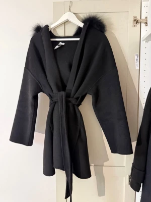 Alicia  kappa - FUR Stockholm - Svart kappa från FUR Stockholm med stor huva kantad av mjuk päls. Modellen har knytband i midjan och vida ärmar för en chill och trendig look. Har inte använt den så mycket. 