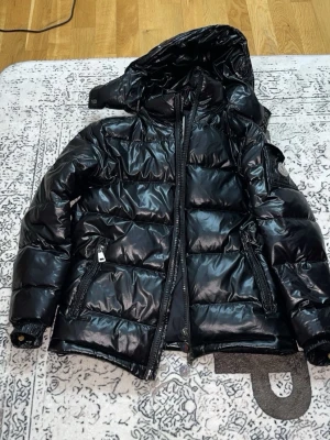 Svart Moncler pufferjacka med huva - Säljer en svart, glansig pufferjacka från Moncler med huva och dragkedja. Jackan har Moncler-logga på ärmen, två fickor med dragkedja och justerbar nederkant. Perfekt för kalla dagar och riktigt snygg streetstil.
