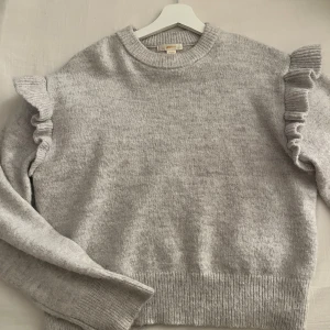 Ljusgrå stickad tröja med volang - Superfin ljusgrå stickad tröja från Gina Tricot med volangdetaljer på axlarna. Tröjan har rund halsringning, långa ärmar och ribbade muddar. Perfekt för dig som vill ha en söt och trendig look till hösten eller vintern. Knappt använd. 