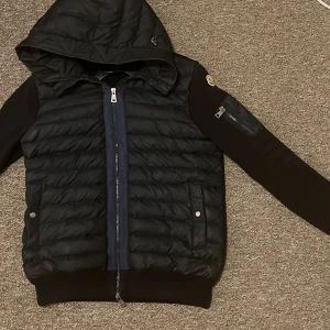 Svart Moncler cardigan med huva - Snygg svart pufferjacka från Moncler med quiltad framsida. en extra ficka med dragkedja på ärmen.Perfekt för dig som vill ha slim jacka som sitter längst kroppen. Size s men passar m” priset kan diskuteras så våga ge förslag