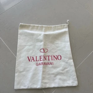 Vit dustbag från Valentino Garavani - Vit dustbag från Valentino Garavani i bomull med röd logga framtill. Påsen har snörning upptill och är perfekt för att förvara din väska eller accessoarer. Enkel och stilren design med klassisk känsla. 