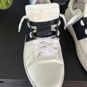 Valentino Garavani open  - (SKO SNÖREN KOMMER OCKSÅ TILL) Snygga vita sneakers från Valentino Garavani med bred marinblå rem över vristen. Skorna har rund tå, platt sula och är tillverkade i skinn. Perfekta för dig som gillar stilrena och exklusiva sneakers med en unik touch.