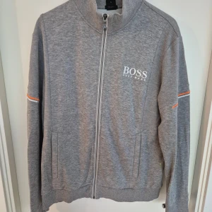 Grå ziptröja från Hugo Boss - Snygg grå tröja från Hugo Boss med dragkedja framtill och vit logga på bröstet. Tröjan har ribbade muddar, diskreta orange detaljer på ärmarna och två fickor framtill. Perfekt för en stilren och sportig look.