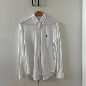Vit skjorta från Polo Ralph Lauren - Vit långärmad skjorta från Polo Ralph Lauren i lätt mesh-bomull. Klassisk button-down krage, knäppning framtill och ikonisk blå broderad logga på bröstet. Perfekt för en clean och stilren look. Skriv för fler bilder och frågor