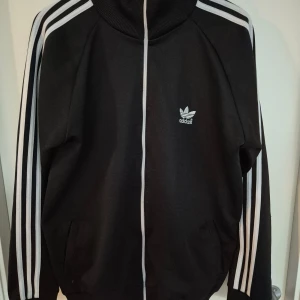 Svart Adidas hoodie med vita ränder - Klassisk svart Adidas hoodie med vita ränder längs ärmarna och dragkedja framtill. Tröjan har hög krage, ribbade muddar och Adidas logga på bröstet. Perfekt för en sportig och avslappnad stil.