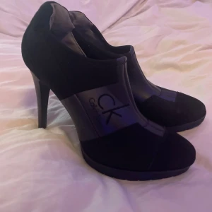 Svarta klackskor Calvin Klein,  - Snygga svarta klackskor från Calvin Klein med höga smala klackar och plattformsula. Skorna har en mix av skinn och textil, med CK-logga på sidan och rund tå. 