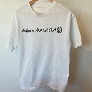 Vit Maison Margiela t-shirt med tryck(nyskick) - Vit t-shirt från Maison Margiela med svart handskrivet logotryck på bröstet. Klassisk rund hals och korta ärmar. Tillverkad i mjuk bomull för en clean och stilren vibe. Perfekt för dig som gillar minimalistisk design med en twist.