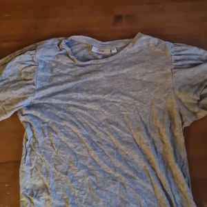 Ljusgrå t-shirt från Zink, storlek S - Ljusgrå t-shirt från Zink i storlek S. T-shirten har korta ärmar med lätt puff vid axlarna och rund halsringning. Tillverkad i mjuk bomull, perfekt för en chill och enkel stil. Liten metallbricka med Zink-logga nertill ger en snygg detalj.